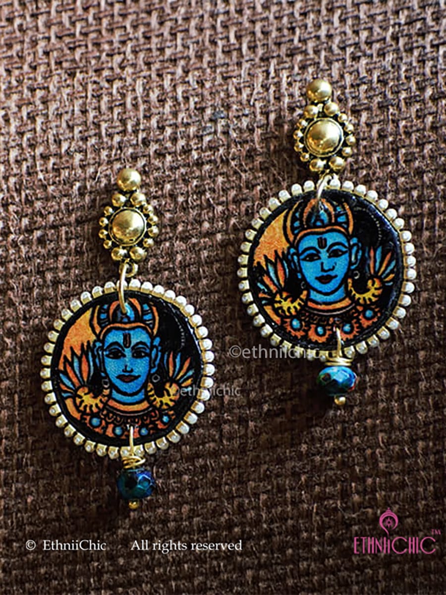 Pair of earrings with a design of Vishnu face small in size hand-painted in Kerala Mural style in a circular frame on a brown fabric background.