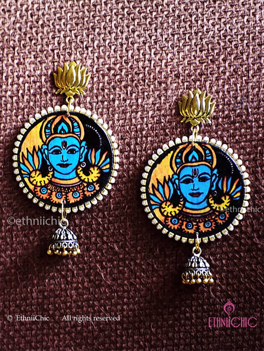 Pair of earrings with a design of Vishnu face hand-painted in Kerala Mural style in a circular frame on a brown fabric background.